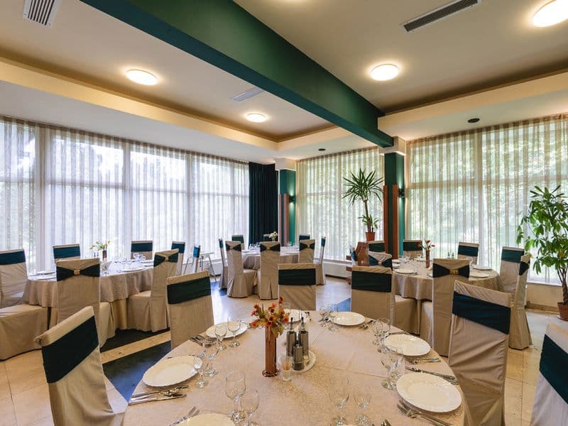 Restaurantul Complexului Bradul-Călimani, cu mese rotunde aranjate lângă ferestre mari cu perdele