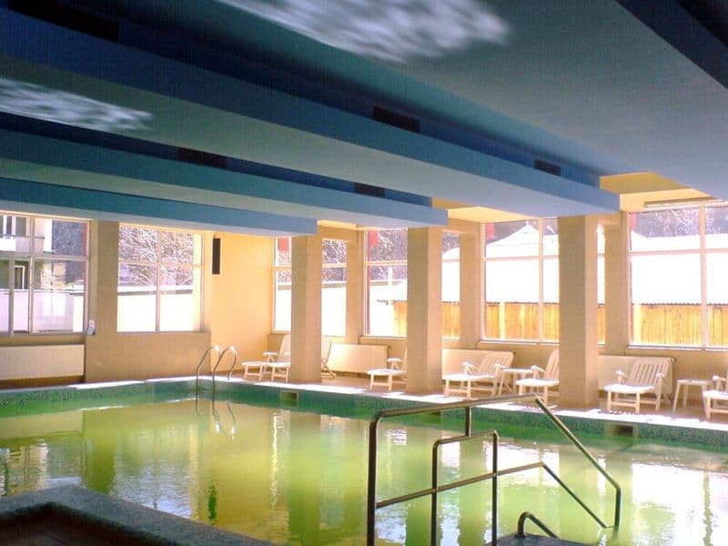 Piscina interioară Bradul-Călimani cu scară de acces, șezlonguri albe și ferestre spre curte